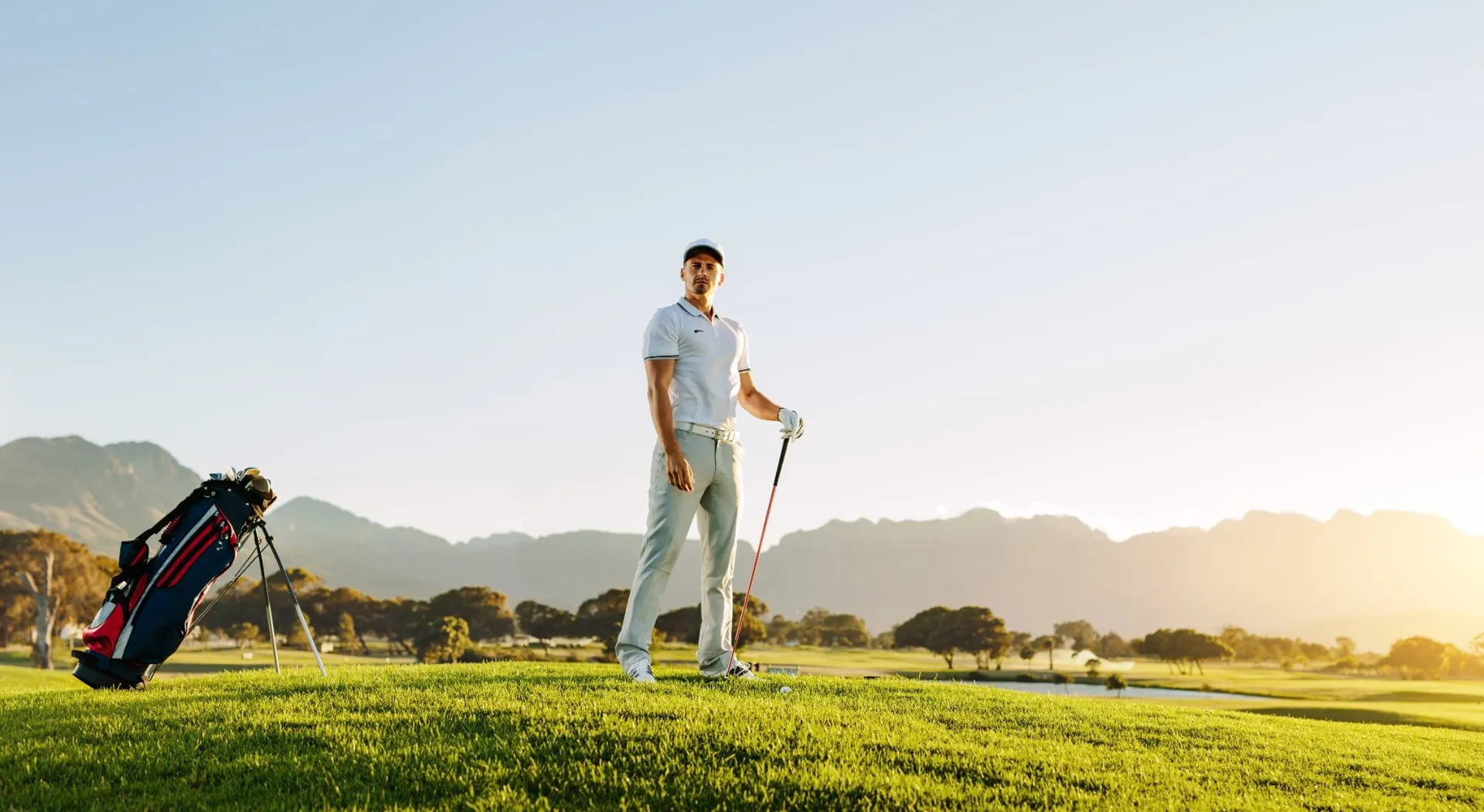 Golfer standing with a golf bag on a sunlit course with mountains in the background